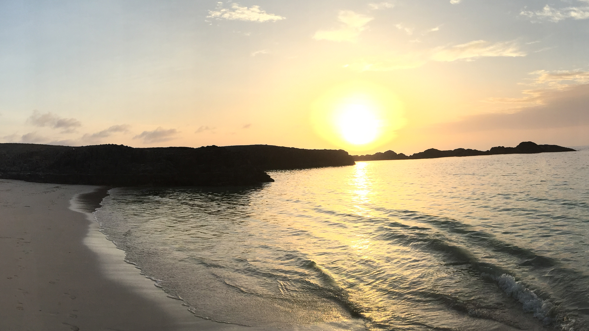 Un vaste coucher de soleil sur une plage au bord d'un océan calme. Des caps rocheux sombres se découpent sur la lumière dorée, qui se reflète sur l'eau et sur le rivage.