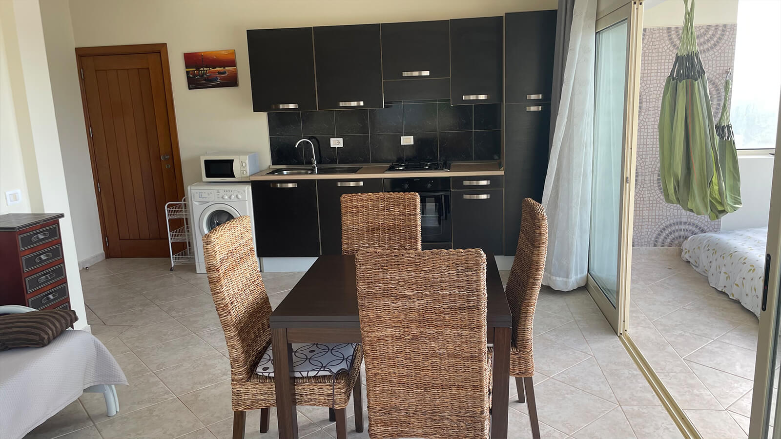 Un coin repas, composé d'une table foncée et de quatre chaises en osier, se trouve face à une kitchenette. La cuisine est équipée de placards foncés, d'un lave-linge et d'un four ; on aperçoit une chaise-hamac à travers la porte ouverte sur la droite.