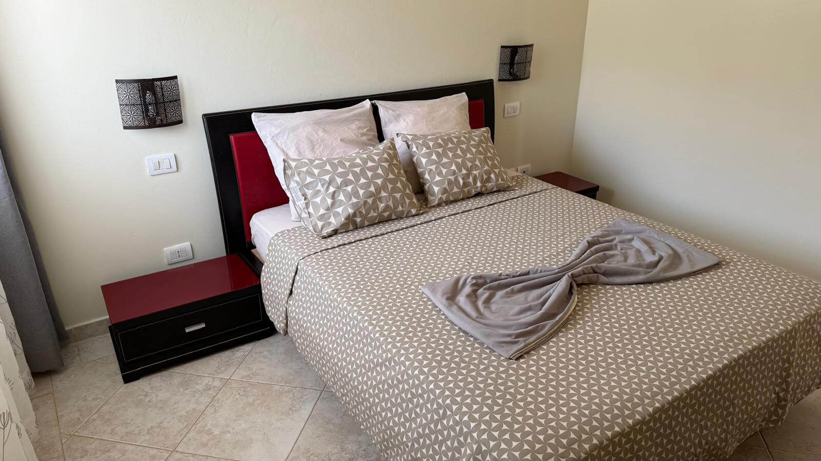 Un lit bien fait, recouvert d'une couette à motifs gris et blancs, de deux coussins décoratifs à motifs et de deux oreillers blancs. Le lit est doté d'une tête de lit noire avec des panneaux latéraux rouges et de tables de chevet noires assorties de chaque côté.